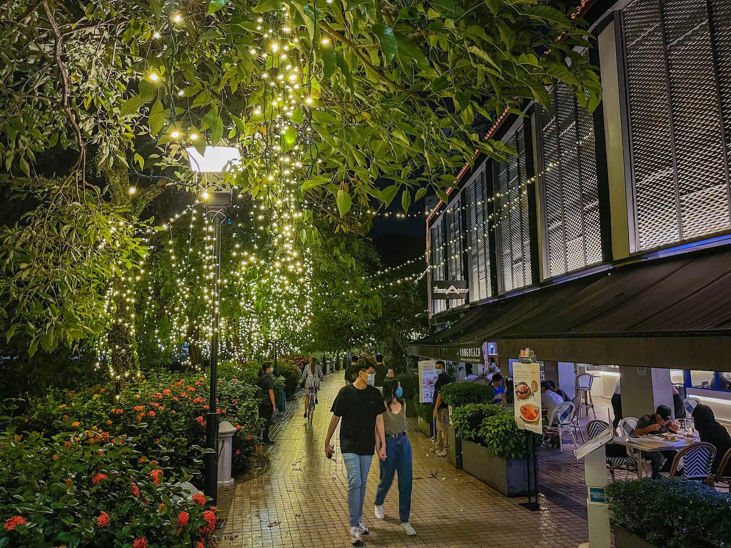 People wearing masks walk a brick path lined with lights and restaurants at night.